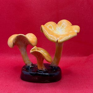May include: A ceramic sculpture featuring three yellow-orange chanterelle mushrooms with visible gills on the undersides of the caps. The mushrooms are attached to a smooth, glossy black oval base, creating a singular decorative object.