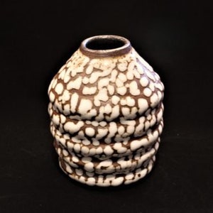 Puede incluir: Un pequeño jarrón de cerámica blanco y marrón con una superficie texturizada y abultada. El jarrón tiene una forma redondeada y una abertura estrecha.