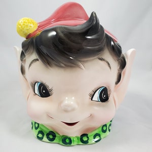 Könnte beinhalten: Ein Vintage-Keksbehälter aus Keramik in Form eines lächelnden Elfenkopfes. Der Elf hat schwarzes Haar, blaue Augen und einen roten Hut mit einem gelben Bommel. Ein grüner Kragen mit dunkelgrünen Akzenten vervollständigt das Design.