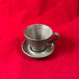 Puede incluir: Un juego de taza y platillo de cerámica gris. La taza tiene una forma redondeada y un asa. El platillo es redondo y tiene un borde ligeramente elevado.