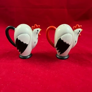 May include: Two ceramic salt and pepper shakers in the shape of white roosters. Each has a black or red handle, black wing detail, and a red comb. The shakers sit on a black base and have a small hole for dispensing.