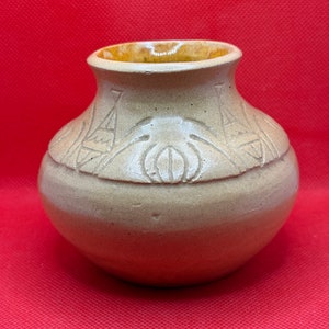 Puede incluir: Un pequeño jarrón de cerámica beige con un cuerpo ligeramente redondeado y una abertura ancha y redonda. El jarrón tiene un diseño decorativo de hojas estilizadas y formas geométricas.