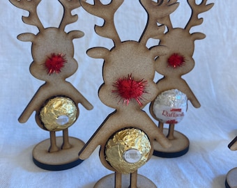 May include: Three wooden reindeer ornaments with red pom-poms for noses and chocolate candies in their bellies. The reindeer are standing on round wooden bases.