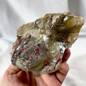 May include: A large, translucent green and brown calcite crystal with red inclusions. The crystal has a rough, natural surface and is irregularly shaped.