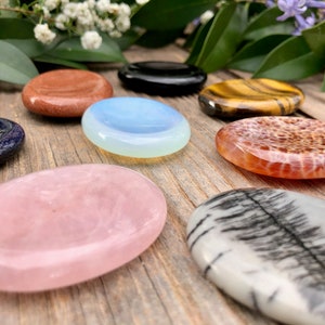 May include: A collection of seven smooth, oval-shaped polished stones in various colors, including pink, blue, white, brown, and black. The stones are arranged on a wooden surface.