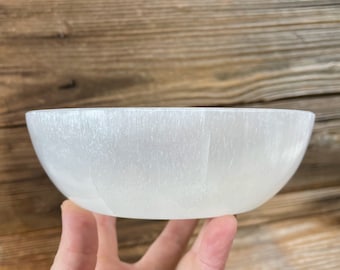 May include: A translucent white bowl crafted from selenite, featuring a smooth, rounded shape and a fibrous, shimmering texture throughout the material. The bowl has a consistent, curved profile with a polished finish that catches the light.
