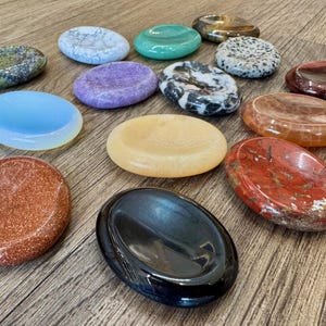 May include: A collection of fifteen polished, oval-shaped stone palm stones and worry stones in various colors and textures, including black, red, orange, yellow, blue, purple, green, and speckled patterns, arranged in a scattered group.