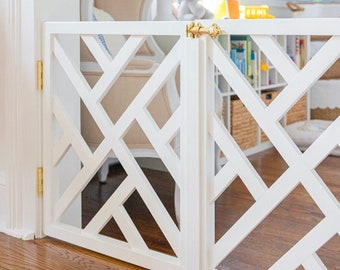 May include: White wooden baby gate with a diamond pattern and gold hinges. The gate is open and is in front of a white chair and a bookshelf.