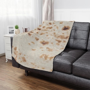 Puede incluir: Una manta de tiro marrón y beige con un patrón que se asemeja a una tortilla. La manta está drapeada sobre un sofá de cuero negro.