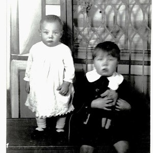 May include: A vintage black and white photograph of two young children. One child stands in a white dress, black shoes, and white socks. The other child sits, wearing a dark outfit, white collar, and holding a doll.