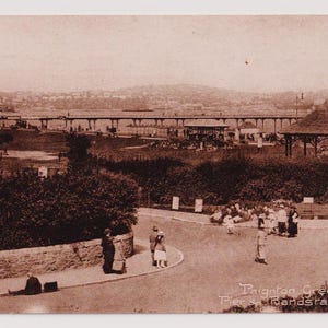 Può includere: Una cartolina vintage in seppia che mostra una vista del molo e del palco per la banda a Paignton Green, in Inghilterra. Il molo è in legno e ha una struttura lunga e stretta. Il palco per la banda è una piccola struttura ottagonale con un tetto. Ci sono persone che camminano sul molo e nel parco.