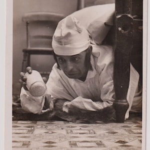May include: A vintage black and white photograph of a man in a nightcap and nightshirt, peering under a bed. He holds a cylindrical object with holes. The image is titled "THE MERRY MERRY HUNTSMAN."