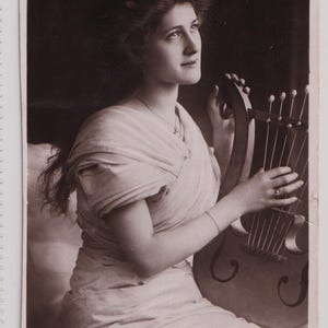 May include: Sepia-toned vintage postcard featuring a woman holding a lyre. She is wearing a draped garment and jewelry. The text on the postcard reads "2009 N ROTARY PHOTO, E.C. MISS PHYLLIS DARE."