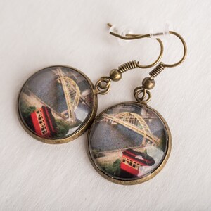 Op de afbeelding: Een paar bronzen oorbellen met een ronde glazen hanger met een vintage afbeelding van een brug en een rode tram.