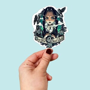 May include: A sticker featuring a cartoon girl with black hair and a black dress, holding a bottle with a skull and crossbones. The sticker has a green and blue background with white clouds and the text "Over Your Dead Body".