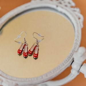 Puede incluir: Un par de pendientes de plata con colgantes de zapatos rojos. Los pendientes se muestran en un espejo circular blanco adornado.