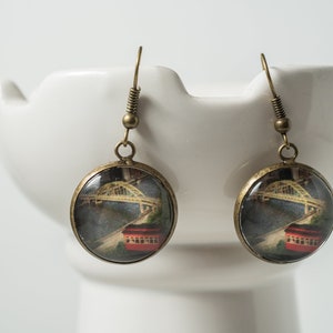 Op de afbeelding: Een paar bronzen oorbellen met ronde glazen cabochons met een vintage afbeelding van een brug en een rode tram.