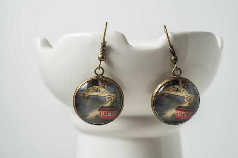 Op de afbeelding: Een paar bronzen oorbellen met ronde glazen cabochons met een vintage afbeelding van een brug en een rode tram.