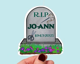 May include: A gray tombstone with the text "R.I.P. JO ANN 1943-2025" and a needle and thread and scissors on it. The tombstone is surrounded by green grass and purple flowers.