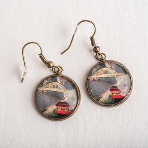 Op de afbeelding: Een paar bronzen oorbellen met ronde glazen cabochons met een foto van een brug en een rood gebouw.