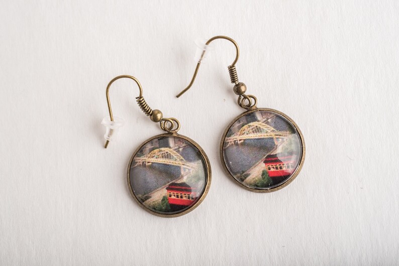 Op de afbeelding: Een paar bronzen oorbellen met ronde glazen cabochons met een foto van een brug en een rood gebouw.