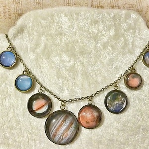 May include: A gold chain necklace with seven round glass pendants featuring images of planets. The planets are in shades of blue, pink, and brown.