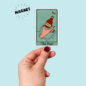 May include: A magnet featuring a tarot card design with a teal background. The card depicts a hand holding a ketchup bottle with the text "Hunt's" on it. The card is labeled "The Fool." The word "MAGNET" is above the card.