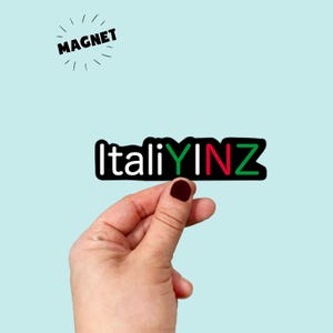 May include: A black magnet with the word "ItaliYINZ" in white, green, and red lettering is held by a hand. The word "MAGNET" is above the magnet in black with radiating lines.