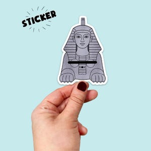 May include: A gray sticker featuring a stylized illustration of the Sphinx. The sticker has a white border and the word "STICKER" in black, with radiating lines. The Sphinx is depicted in a seated position.