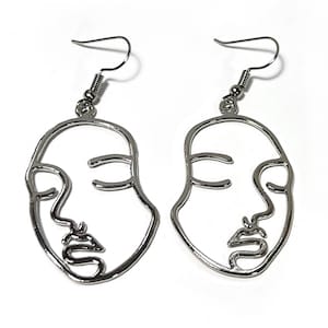 May include: A pair of silver earrings with a line art design of a face. The earrings are dangle style with a hook closure.