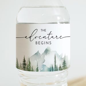 Könnte beinhalten: Eine transparente Wasserflasche mit einem Etikett mit der Aufschrift "The Adventure Begins". Das Etikett zeigt ein Aquarelldesign von Bergen und Bäumen in Grün und Blau. Die Flasche ist mit klarer Flüssigkeit gefüllt.