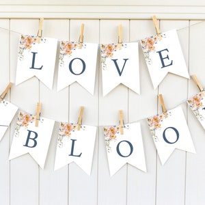 May include: A white banner with floral accents and the text "LOVE in BLOOM" in navy blue lettering. The banner is strung with twine and wooden clothespins.
