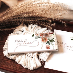 May include: A white chocolate bar wrapper with a floral design and the text "Fall in love". The wrapper is on a white crocheted coaster and a wooden tray.