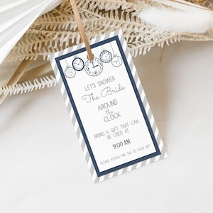 Puede incluir: Una etiqueta rayada gris y azul sobre un fondo blanco. La etiqueta tiene un tema de reloj con varios relojes colgando de la parte superior. El texto en la etiqueta dice "Let's shower the bride around the clock. Bring a gift that can be used at 9:00 AM. Please attach this tag to your gift."