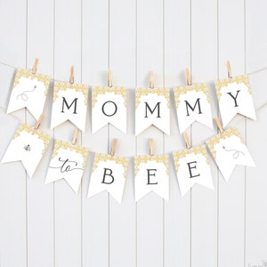 Puede incluir: Bandera blanca con un patrón de panal de abeja amarillo y texto negro que dice "Mommy to Bee". La bandera está atada con cuerda y pinzas para la ropa.