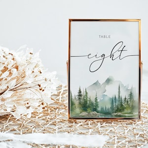 Puede incluir: Un cartel de número de mesa con marco dorado con una ilustración de acuarela de una cordillera y un bosque. El cartel dice "TABLE eight".