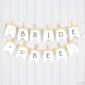May include: White and yellow bridal shower banner with the text "Bride to Bee" in black lettering. The banner is decorated with a honeycomb pattern and small bees.