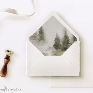 May include: White envelope with a watercolor design of a foggy forest scene on the flap. The envelope is lying flat on a white surface with a wax seal and a white ribbon.