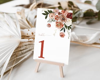 May include: A white table number card with a floral design in shades of red, pink, and green. The card is on a small wooden easel and the number "1" is written in red. The text "table" is written in cursive above the number.