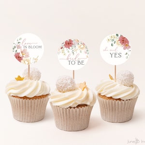Może przedstawiać: Trzy babeczki z kwiatowymi ozdobami. Ozdoby mówią "love in bloom", "bride to be" i "she said yes".