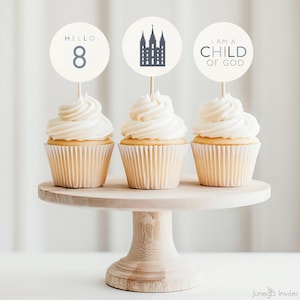 May include: Three cupcakes with white frosting and decorative toppers on a wooden cake stand. The toppers read "HELLO 8", a church illustration, and "I AM A CHILD OF GOD". The cupcakes are in beige paper cups.
