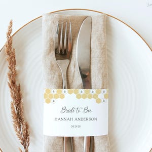 Pode incluir: Um cartão de lugar branco com um padrão de favo de mel amarelo e o texto "Bride to Bee" está amarrado em torno de um guardanapo de linho com um garfo e uma faca. O cartão de lugar também inclui o nome "Hannah Anderson" e a data "04.29.28".