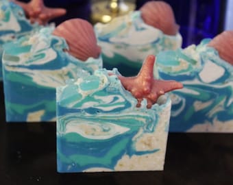 May include: Four handmade soap bars with a beach theme. Each bar is decorated with a seashell and a starfish. The soap bars are blue, white, and green.