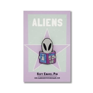 Puede incluir: Un pin de esmalte suave con un extraterrestre gris leyendo un libro titulado "Aliens". La portada del libro es rosa y azul con un dibujo de una nave espacial extraterrestre. El pin está sobre un fondo blanco con una estrella morada.