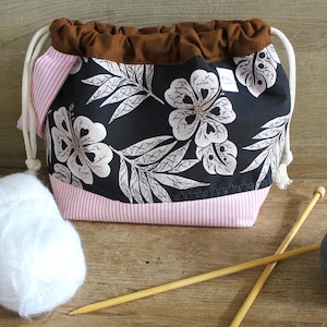 Peut inclure: Un sac à cordon noir avec un fond rayé rose et blanc et un imprimé floral représentant des fleurs d'hibiscus blanches et des feuilles sur un fond noir. Le sac est rempli de fil blanc et de deux aiguilles à tricoter en bois. Une petite plante succulente en pot se trouve au premier plan.