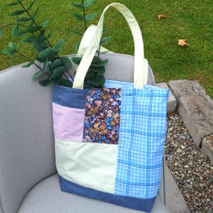 May include: A patchwork tote bag featuring a combination of fabrics including light blue plaid, a dark floral print, light pink and white stripes, solid pale yellow, and a dark blue denim base with two pale yellow fabric handles.