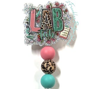 May include: A retractable badge reel with a glittery "LAB tech" design. The design includes pink, green, and leopard print elements. The reel has a pink, leopard print, and teal bead accent, and a clear plastic clip with a pink flower.