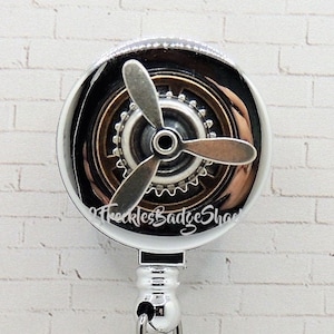 May include: A silver retractable badge reel with a propeller design and a small airplane charm. The reel has a round face with a propeller and gear details. The airplane charm hangs below on a silver chain.
