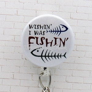 May include: White round button with a black and white fish skeleton design and the text "Wishin' I Was Fishin'". The button is attached to a silver retractable badge reel with a silver fish skeleton charm.