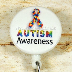 May include: White button with a blue, red, yellow, and green puzzle piece ribbon and the text "Autism Awareness". A matching puzzle piece ribbon charm is attached to a retractable badge holder.